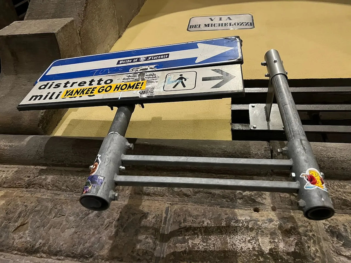 A metal pedestrian crossing arm with a blue-and-white directional sign and a pedestrian symbol, photographed outdoors against a stone wall and a yellow wall in Florence, Italy.