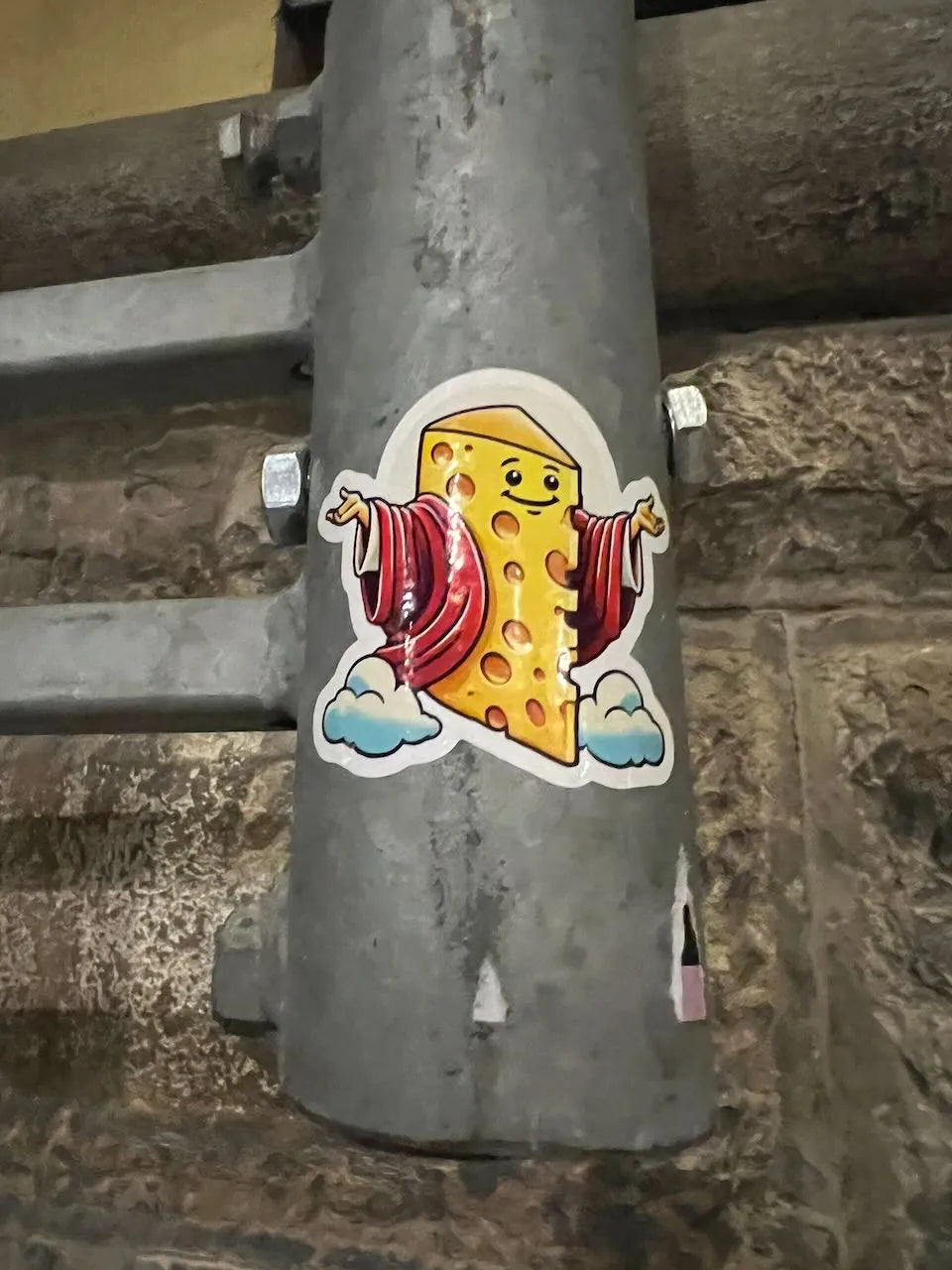 A die-cut sticker of a smiling cheese slice wearing a red robe and blue clouds, applied to a gray metal pipe outdoors against a stone wall in Florence, Italy.