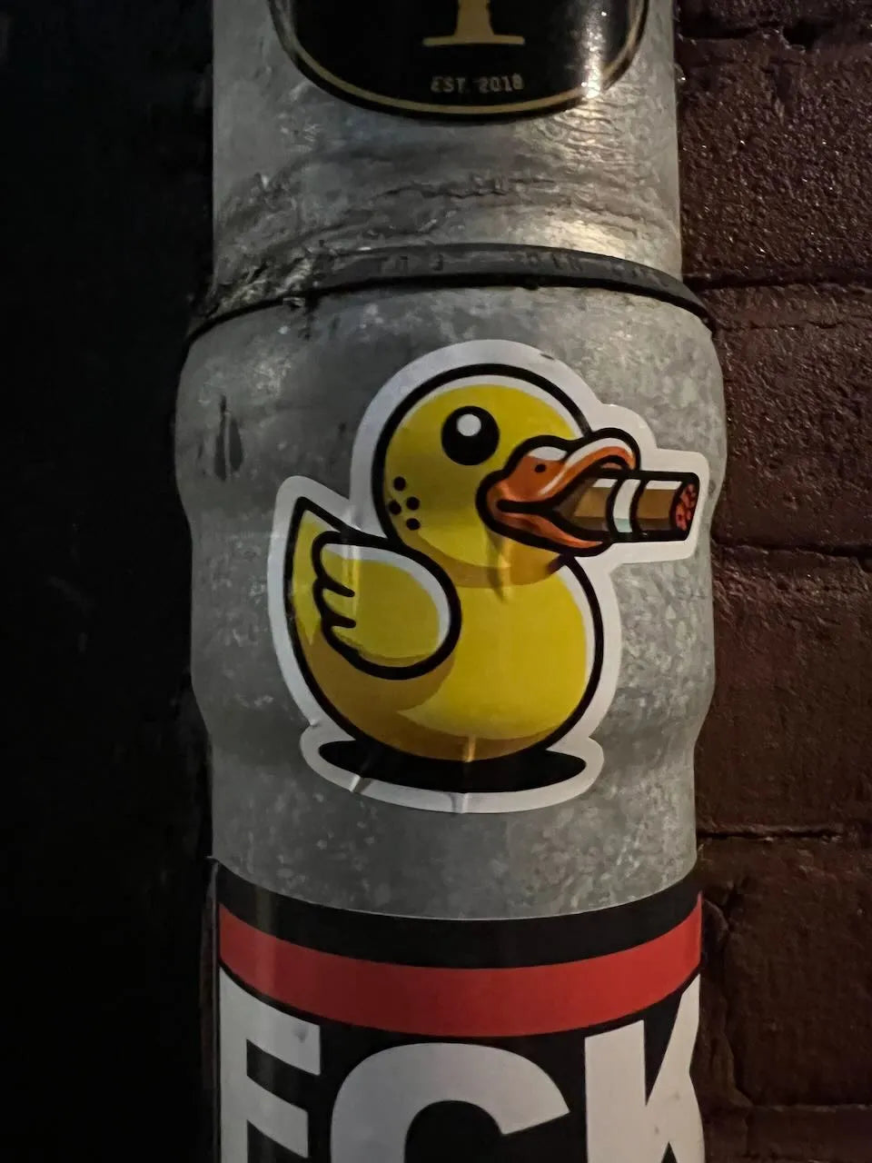 A close-up of a yellow rubber duck sticker on the front, featuring a stylized duck holding a cigar, with partial black, red, and white branding text visible at the bottom.