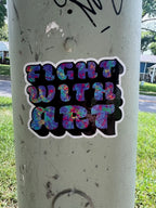 A die-cut vinyl sticker with the text 'FIGHT WITH ART' in a colorful, glittery multicolor font with a black background and white border, shown applied to a concrete utility pole outdoors.