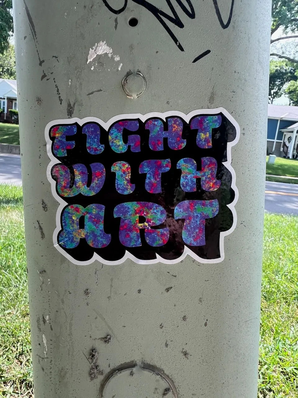 A die-cut vinyl sticker with the text 'FIGHT WITH ART' in a colorful, glittery multicolor font with a black background and white border, shown applied to a concrete utility pole outdoors.