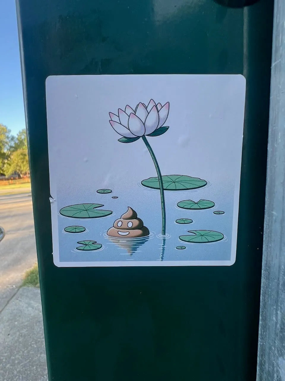 A square sticker with a cartoon illustration of a white lotus flower, green lily pads, and a smiling brown poop emoji floating in water, shown applied to a dark green outdoor box.
