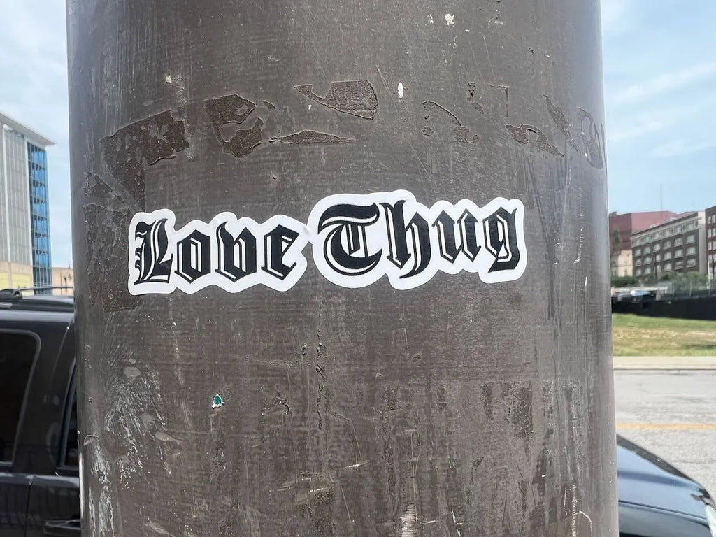 A die-cut vinyl sticker with the text 'Love Thug' in a black gothic font with a white outline, shown applied to the side of a dark metal utility pole in an outdoor urban setting.
