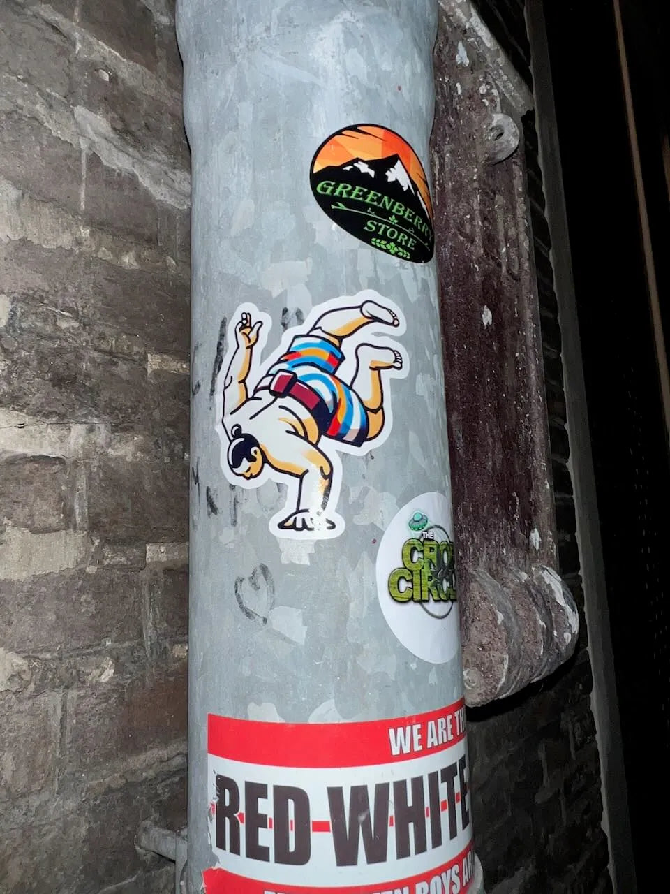 A close-up of a gray metal utility pole covered with stickers, including a sumo b-boy sticker set against a dark brick wall.